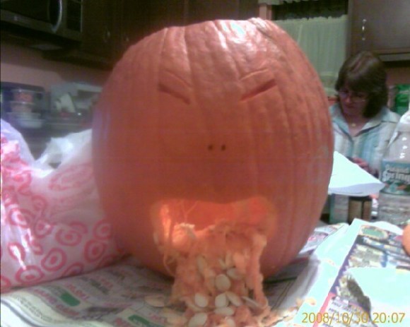 A pumpkins sits on newspapers spread on a table. Slimy pumpkin innards exude from the unhappy-looking pumpkin’s mouth.