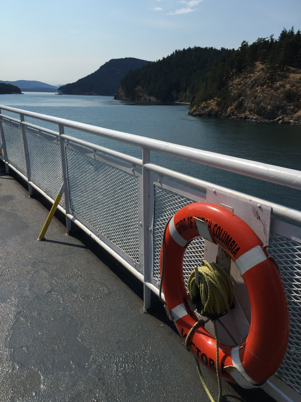 Beyond the deck of a ferry, tree-lined islands are seen in the Strait of Georgia. A life raft attached to the ship's railing gives the name of the ferry, The Spirit of British Columbia.