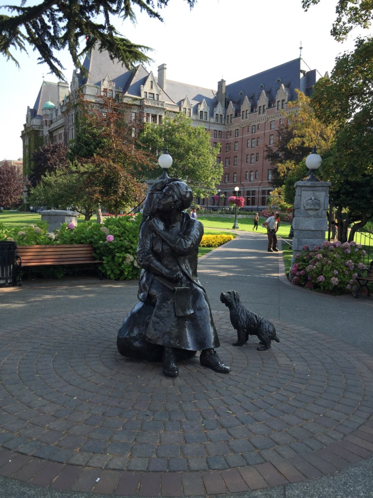 Statue of Emily car seated, with her dog, monkey, and notebook. The Fairmont Empress Hotel stands behind the statue.